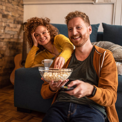 Man and woman smiling and watching TV in their home while eating popcorn