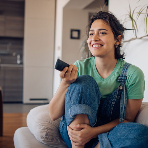 Woman sitting on a couch smiling while holding a remote and watching tv